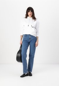 Sudadera blanca con cordones ajustables, jeans rectos azules y zapatos negros. Sosteniendo un estuche de guitarra negro. Fondo simple.