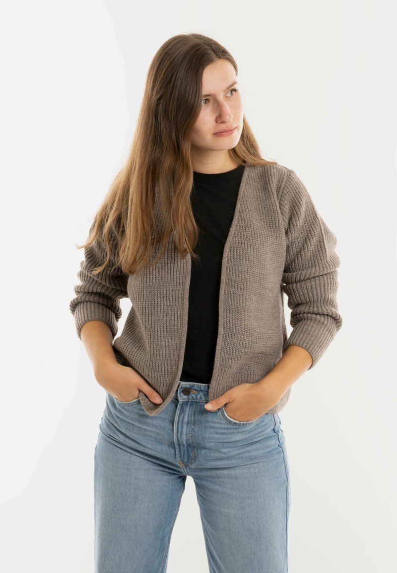 Gebreide cardigan in lichtbruin met geribbelde textuur en open voorkant; gedragen over een zwarte top, gecombineerd met blauwe jeans.