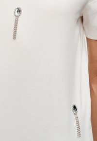 Tissu blanc orné de deux chaînes argentées et de pierres précieuses, l'une près de l'encolure et l'autre près de la hanche.