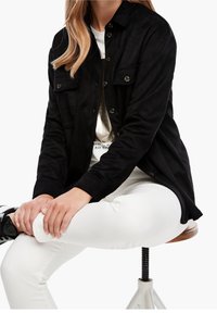 Donna seduta su uno sgabello, con indosso una giacca nera con bottoni, pantaloni bianchi, una camicia bianca con stampa metallica e scarpe nere.