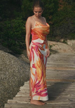 Junge Frau steht barfuß auf einem Holzweg, trägt ein langes Kleid mit orange-pinkem Blumenmuster und schaut nach unten.