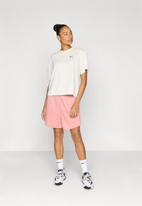 Ανοιχτό κρεμ κοντομάνικο τοπ με μαύρο λογότυπο, συνδυασμένο με ροζ αθλητικό σορτς. Το μοντέλο φοράει λευκά αθλητικά παπούτσια και κάλτσες με λογότυπο.