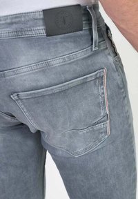 Jean en denim gris clair avec une poche arrière, des coutures visibles et une étiquette en cuir noir au niveau de la taille avec un logo circulaire.