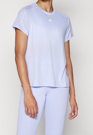 Vrouw draagt lichtpaarse Adidas shirt met korte mouwen en bijpassende legging, staand tegen een effen witte achtergrond.