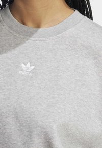 Ljust grå sweatshirt med rund halsringning, tillverkad av mjukt tyg. Har en liten vit Adidas-logotyp på vänster bröst.
