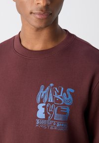 Jeune personne portant un sweat-shirt bordeaux avec un texte graphique bleu « Mind's Eye Scotch & Soda Amsterdam » sur la poitrine.