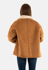 Bruine suede jas met een crèmekleurige schapenvachtkraag, oversized pasvorm en strakke lijnen. De achteraanzicht toont naaddetails en een afgeronde zoom.