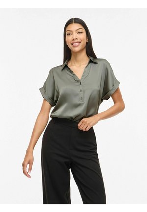 Femme souriante, portant un chemisier en satin vert olive à manches courtes, rentré dans un pantalon noir taille haute, debout avec une main sur la hanche.