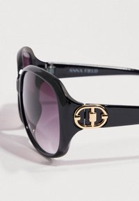 Μαύρα γυαλιά ηλίου με γυαλιστερό φινίρισμα, φακούς με βαθμιαία απόχρωση μοβ και χρυσή διακοσμητική λεπτομέρεια λογότυπου στο πλάι. Μεγάλο, στρογγυλεμένο σχήμα σκελετού.