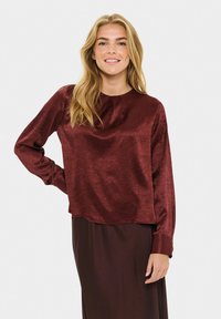 Nasmejana ženska z dolgimi blond lasmi nosi burgundski satenski dolg rokav bluzo in ujemajočo krilo, pozira ob enobarvni beli ozadju.
