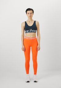 Nike Pro sportsbh i marine med utskjæring, paret med klare oransje leggings med Nike-logo. Materialet er elastisk; designet er tettsittende.