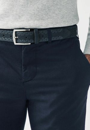 Pantalon bleu marine avec une texture lisse, associé à une ceinture tressée sombre avec une boucle rectangulaire en argent et des trous perforés.