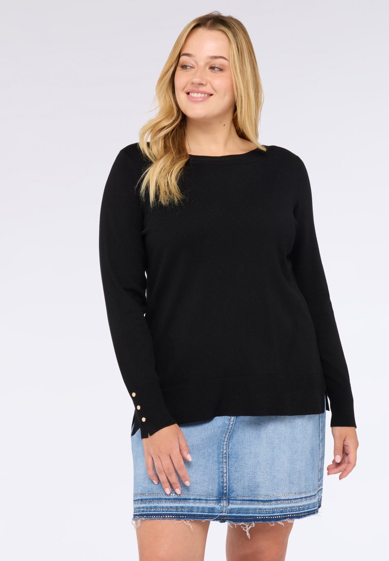 Glimlachende vrouw met lang blond haar, gekleed in een zwarte trui met knoopdetails op de mouwen en een blauwe denimrok met rafelige zoom.