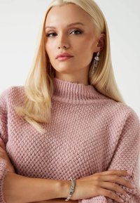 Svetlo roza pletena majica z visokim, strukturiranim ovratnikom. Model nosi dolge, sijoče uhane in srebrno zapestnico na zapestju.