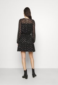 Vestido negro con mangas largas transparentes, salpicado de acentos en oro rosa. Presenta una cintura entallada y una falda acampanada. Un detalle de espalda descubierta.