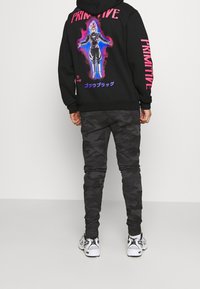 Černá mikina s kapucí s růžovým nápisem „PRIMITIVE“, na zádech barevná grafika a maskáčové kalhoty. Outfit doplňují bílé tenisky.