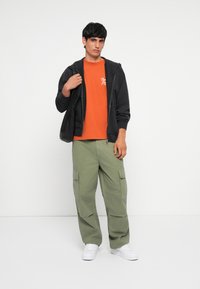 Sort lynlåshættejakke, orange t-shirt med hvidt print, olivengrøn cargo-bukser, hvide sneakers. Model står foran en ensfarvet baggrund.