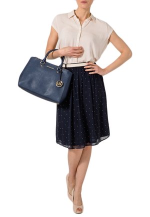 Jupe marine à pois, blouse beige, tenant un sac à main en cuir bleu foncé avec des accents dorés et un logo, associé à des escarpins nus.