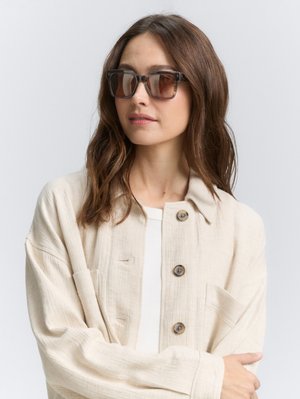 Des lunettes de soleil teintées marron avec une monture noire épaisse et angulaire. Le modèle porte une veste texturée beige clair sur un haut blanc.