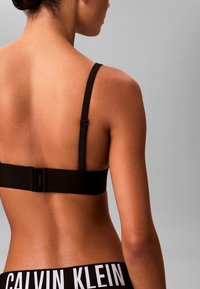 Zwarte beha met verstelbare banden, voorzien van een gladde textuur en een haaksluiting op de rug. Bijpassende zwarte onderbroek met een witte logoband.