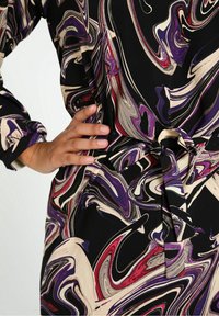 Ruka spočívající na pase, oblečená v šatech s vícivrstvým vzorem ve fialové, červené, černé a béžové barvě, s uvázaným páskem.