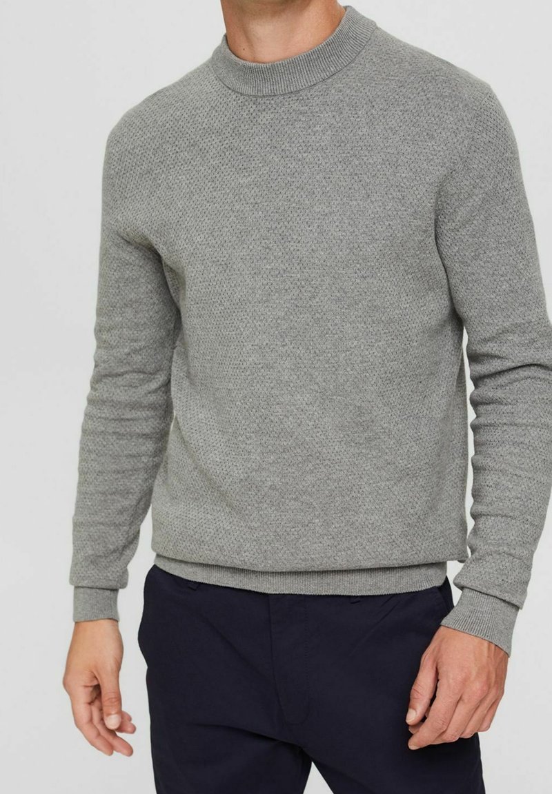 Šedý pletený svetr s kulatým výstřihem, texturovaným vzorem, žebrovanými manžetami a pasem. Nosený přes tmavé kalhoty, látka vypadá měkce.