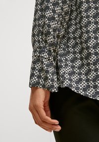 Long-sleeve shirt in navy blue fabric with off-white abstract pattern. Buttoned cuff detail and smooth texture. Worn over black pants.