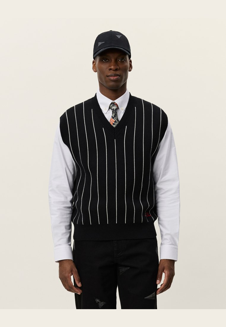 Svart strikket vest med hvite vertikale striper, V-halsdesign, båret over en hvit skjorte med et blomsterbånd, komplett med en svart caps.