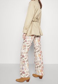 Béžový trench coat s páskem a rozšířeným lemem, spárovaný se světlými kalhotami s květinovým vzorem a hnědými semišovými kotníkovými botami.