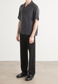 Camisa de manga corta, con botones, en tejido gris oscuro; corte holgado, cuello extendido y pantalones negros lisos complementados con mocasines negros.