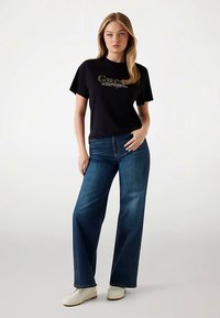 Schwarzes T-Shirt mit kurzen Ärmeln und Logo, kombiniert mit blauen Jeans mit weitem Bein und weißen Sneakern. Glatter Stoff und maßgeschneiderte Passform.