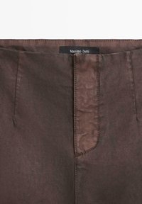 Braune Hose aus Baumwollmischung mit glatter Textur, erkennbarer Naht und einem Etikett mit der Aufschrift "Massimo Dutti" in der Nähe der Taille.