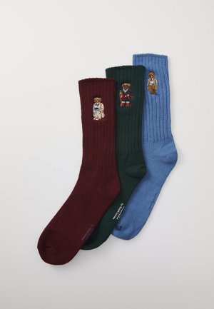 Drei gerippte Socken in Burgunderrot, Dunkelgrün und Blau mit gestickten Teddy-Bären, die auf jeder Socke unterschiedliche Outfits tragen.