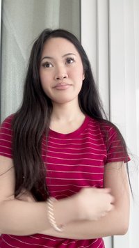 Young woman with long dark hair and red striped shirt, arms crossed, looking thoughtfully to the side indoors by curtains and window.