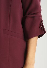 Burgundské sako s texturovanou tkaninou, s rolovanými rukávy s knoflíkovým akcentem a jemným detailním kapsou na přední části.