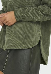 Ręce złożone na mankiecie żakietu w oliwkowo-zielonym kolorze z guzikami, w połączeniu z ciemnozieloną skórzaną spódnicą z przednim srebrnym zamkiem. 