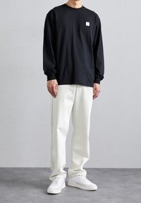 Sort langærmet t-shirt med hvidt logo, parret med hvide denimbukser og hvide sneakers, mod en neutral grå baggrund.