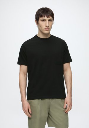 Jeune homme aux cheveux courts et foncés, portant un t-shirt noir uni à manches courtes et un short vert clair sur fond blanc.