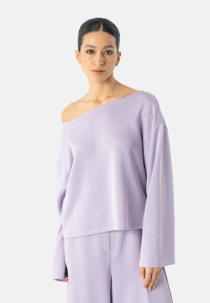 Kobieta z ciemnymi włosami związanymi do tyłu nosi luźną lawendową bluzkę z odkrytymi ramionami oraz pasujące szerokie spodnie na jednolitym tle.
