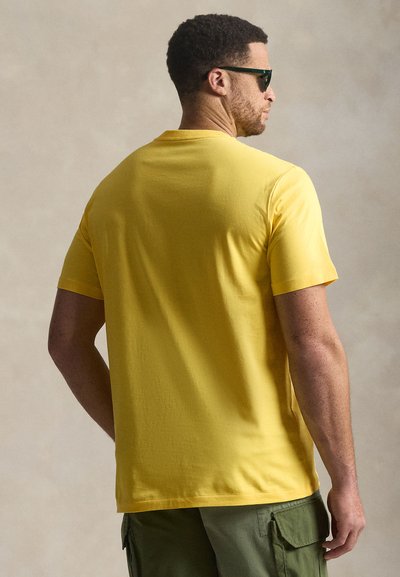 Κοντομάνικη κίτρινη μπλούζα με λείο ύφασμα και στρογγυλή λαιμόκοψη, φορεμένη με παντελόνι cargo, ορατή από πίσω.