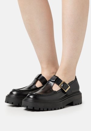 Schwarze Lacklederschuhe mit einer dicken, schwarzen Gummisohle, die ausgeschnittene Seiten und einen goldenen Schnallenakzent aufweisen.