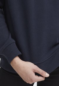 Tamno plavi dukserica s rebrastim manžetama i donjim rubom, meke teksture i opuštenog kroja. Ruka drži donji rub odjeće.