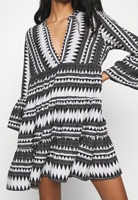 Svart och vit mönstrad klänning med djup V-ringning, långa ärmar och volangkant. Har geometriska zick-zack och triangulära mönster.