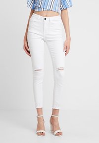 Vaqueros de denim blanco con un corte ajustado, desgastes en las rodillas, dobladillo deshilachado y cintura alta. Combinados con sandalias de tacón blanco.
