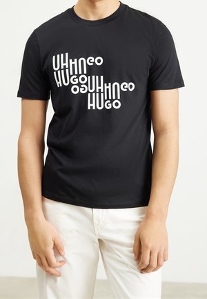 Person trägt schwarzes Kurzarm-T-Shirt mit weiß verzerrtem Text "HUGO", der diagonal wiederholt wird, und weiße Hose vor einfachem Hintergrund.