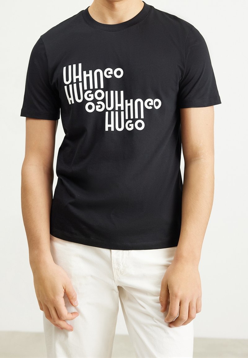 Personne portant un T-shirt noir à manches courtes avec un texte blanc déformé "HUGO" répété en diagonale et un pantalon blanc sur un fond uni.