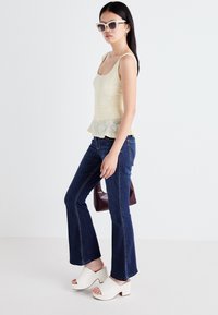 Een vrouw draagt een lichtgele tanktop en een flared jeans, gecombineerd met witte platformklompen en oversized zonnebrillen, staande voor een eenvoudige achtergrond.