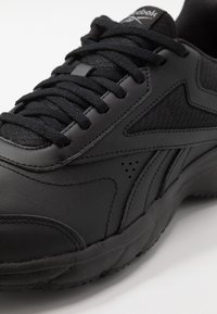 Zapato deportivo negro con exterior de cuero liso, panel de malla texturizada, cordones planos y detalles sutiles de perforación cerca de la punta.
