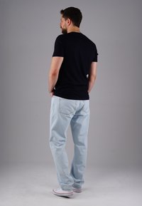 Mann mit Bart, der ein schwarzes T-Shirt, hellblaue Jeans und weiße Turnschuhe trägt, mit den Händen in den Taschen steht und sich von der grauen Hintergrund abwendet.