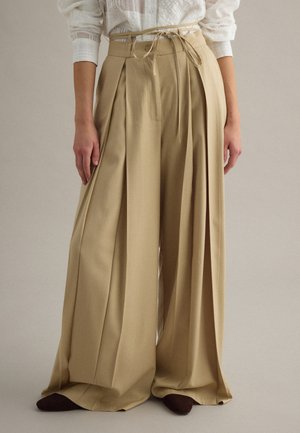 Pantalones beige de pierna ancha con pliegues profundos y un cinturón delgado, combinados con una blusa blanca texturizada y zapatos oscuros visibles en el dobladillo.